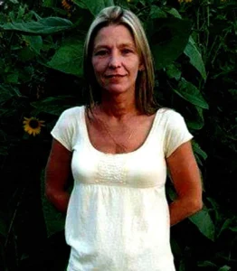 A woman stands in front of tall sunflowers, wearing a white shirt and blue jeans, with her arms relaxed at her sides.