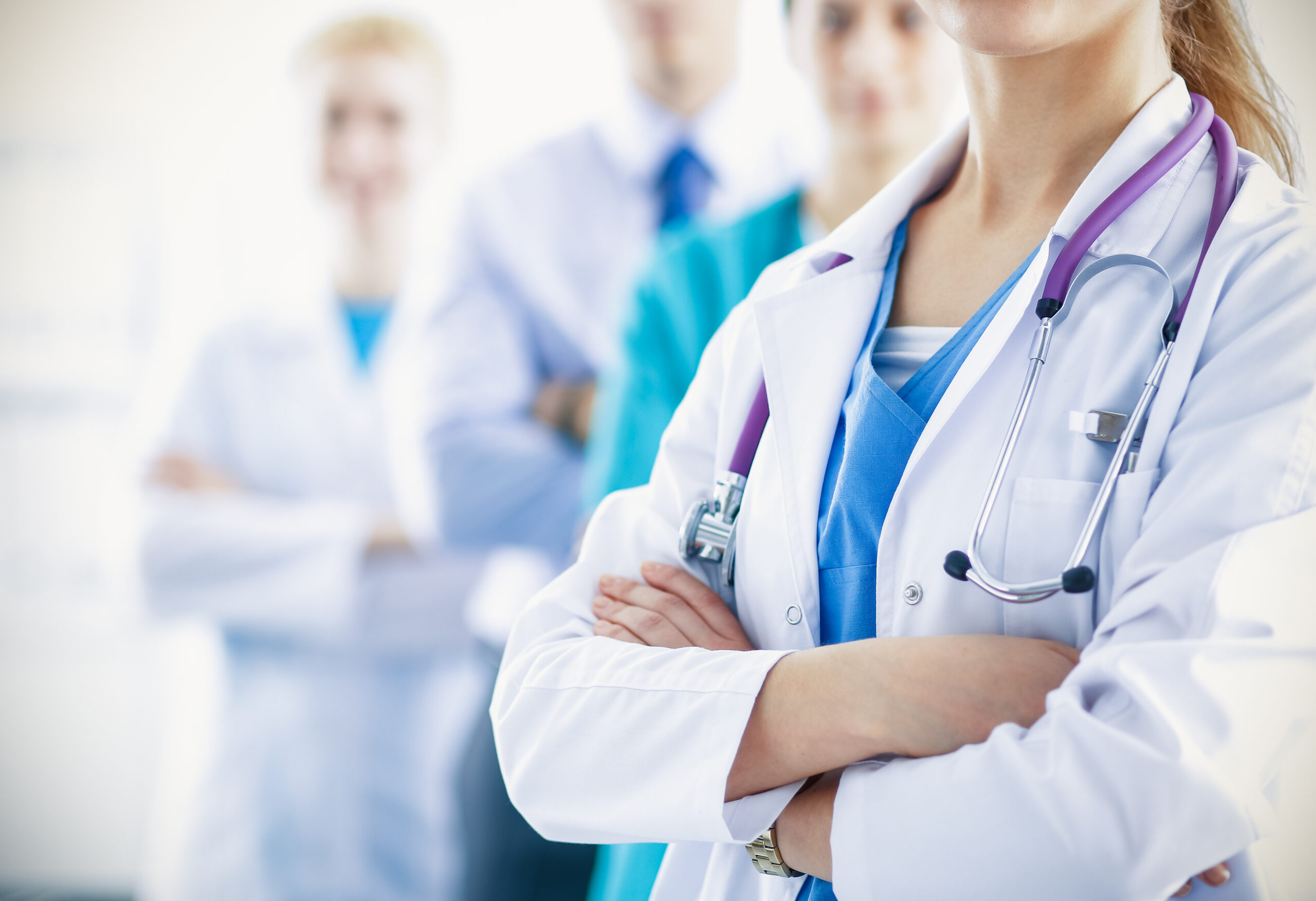A healthcare professional in a white coat with a stethoscope stands confidently, arms crossed, with colleagues blurred in the background.